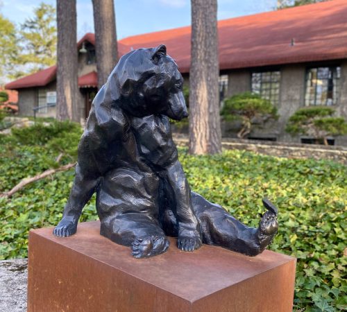 Bronze bear sculpture by artist Roger Martin sitting on a base outside in a garden.