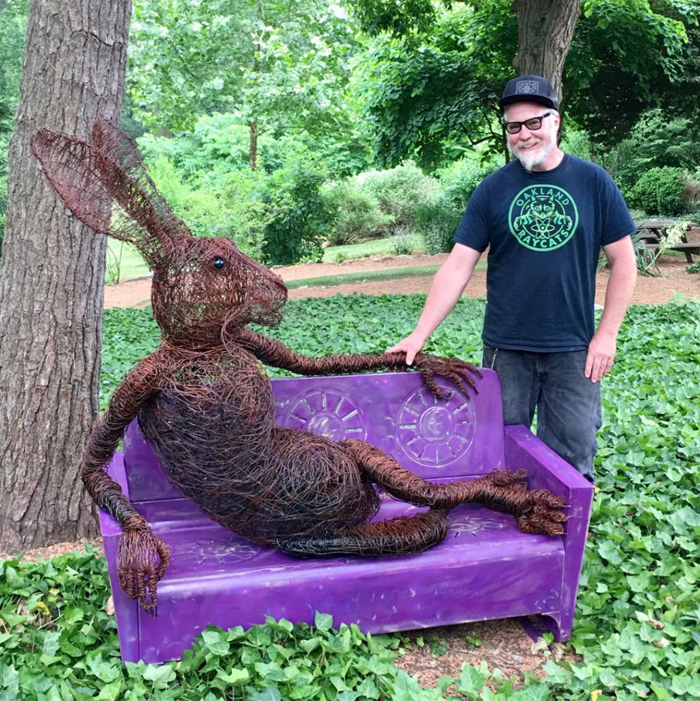 Artist Josh Cote outside in the sculpture garden at Grovewood Gallery posing next to one of his wire hare sculptures.