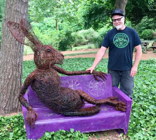 Artist Josh Cote outside in the sculpture garden at Grovewood Gallery posing next to one of his wire hare sculptures.