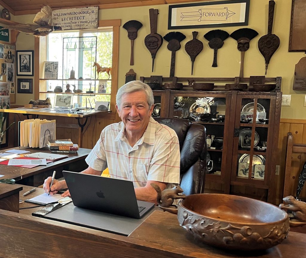 Asheville historian Bruce Johnson sitting at the laptop in his home office surrounded by examples of Biltmore Industries woodcarvings.