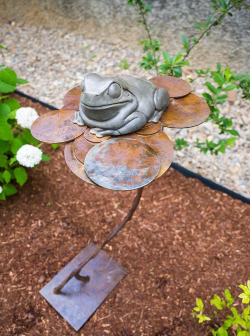 An outdoor sculpture featuring a bronze frog on a steel lily pad titled Fly Fishing by Todd Frahm.