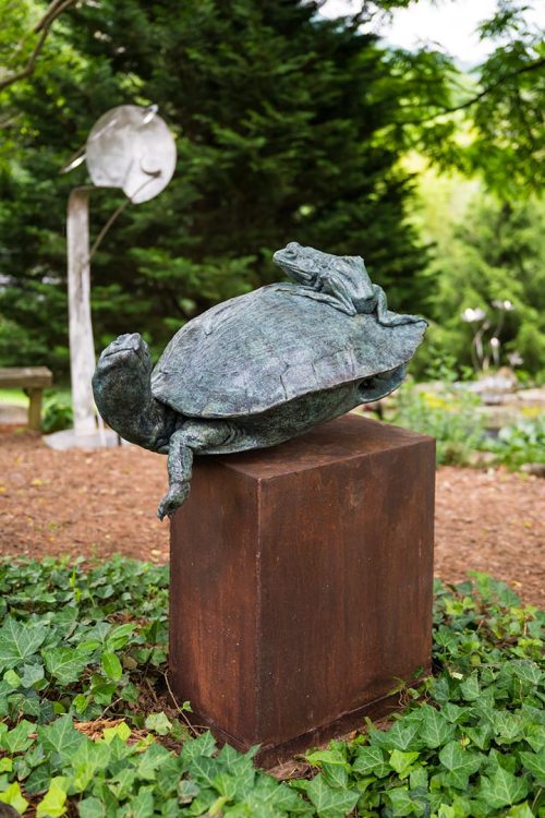 An outdoor sculpture titled The Interloper featuring a large turtle with a frog riding on his back.